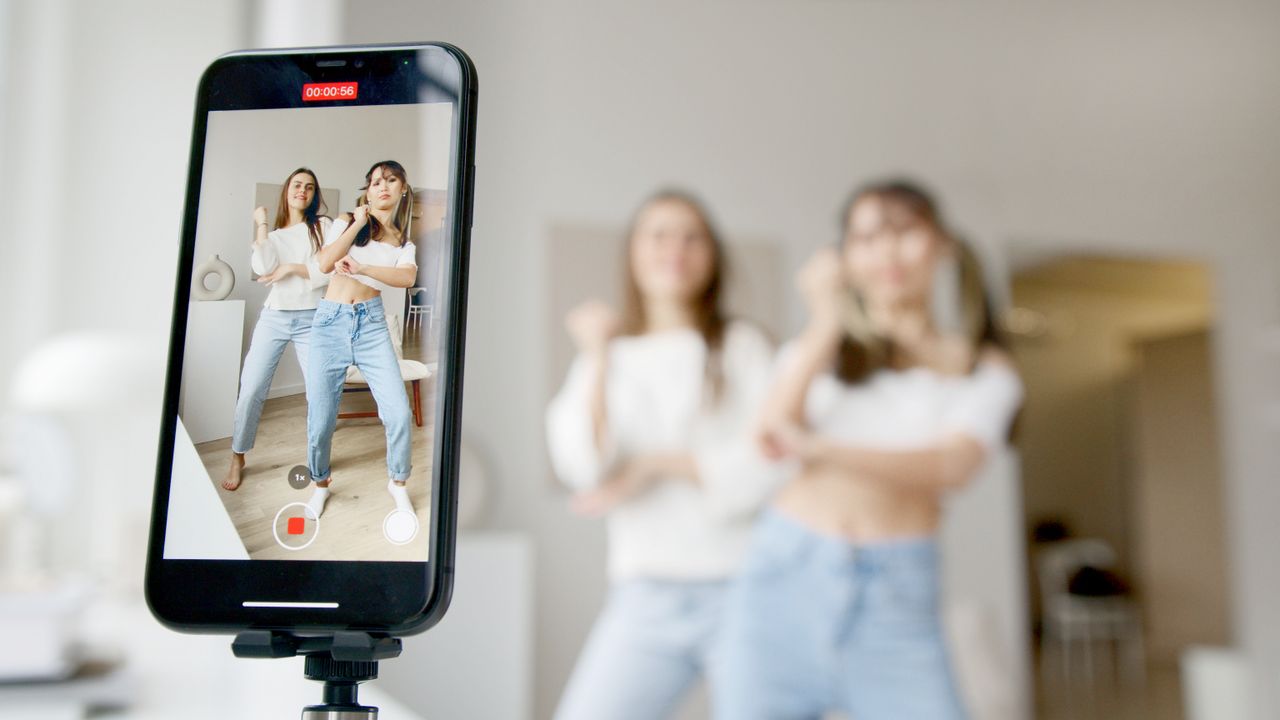 about-us-02 Two women dance in front of a smartphone camera, recording content indoors.